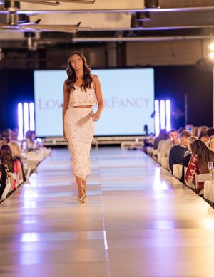 Models walk down a brightly lit white catwalk displaying formal dresses at an indoor fashion event.