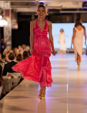 Model walks runway in flowing hot pink dress during fashion show with dramatic lighting and white catwalk.