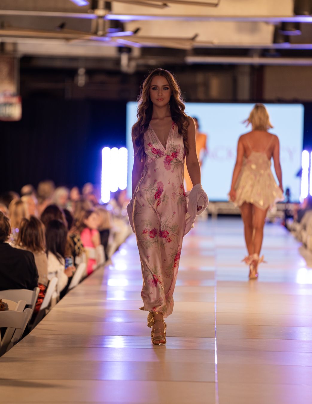 Model walks runway in floral pink sheer maxi dress during fashion show with audience seated along catwalk.