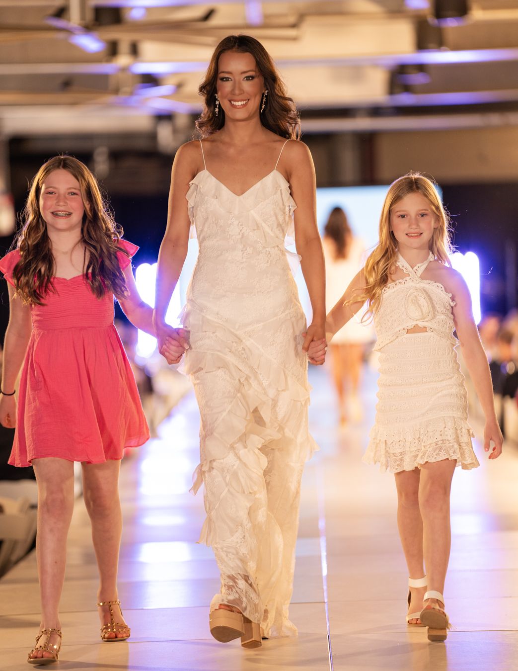 Three models walk the runway together at a fashion show, with the center wearing a flowing white gown and two in shorter dresses.