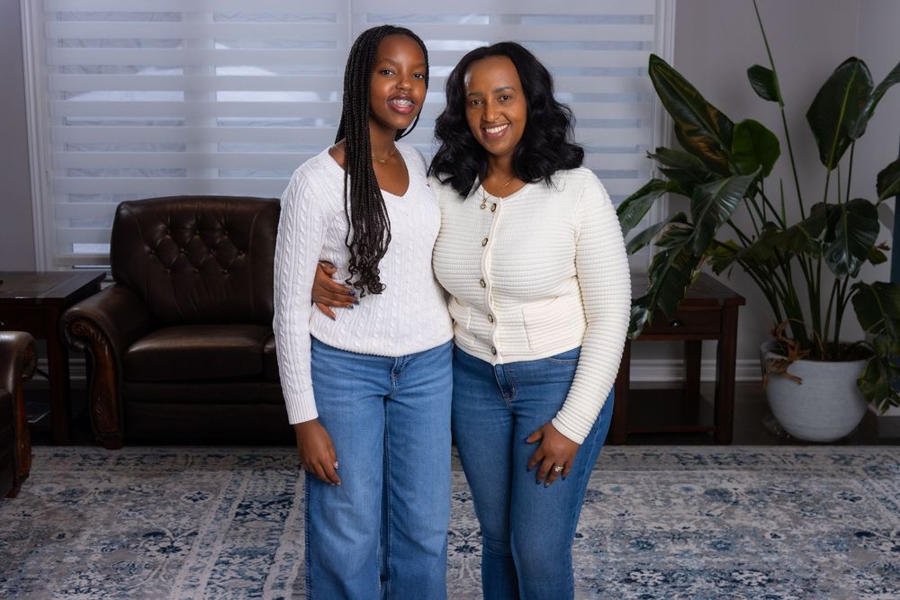 Two people in blue jeans and white tops pose together in front of a window with indoor plants.