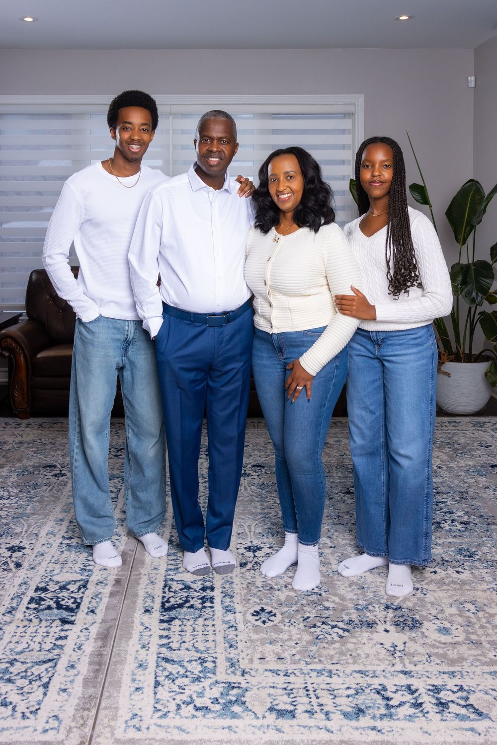 A family wearing matching white tops and blue jeans poses together on a patterned rug in a bright interior space.