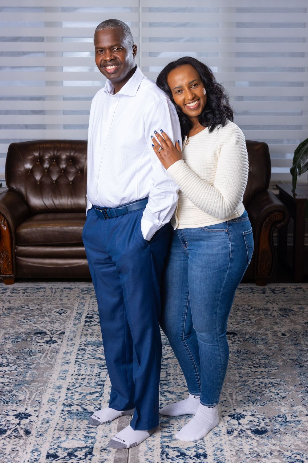 A couple wearing coordinated white sweaters and blue jeans pose together on a patterned rug in a living room.