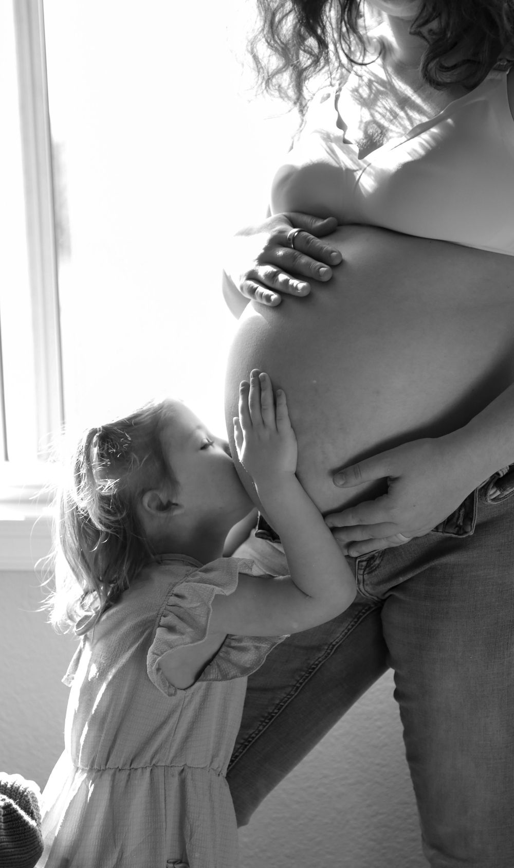 Young child kissing the belly of her pregnant mother i front of bedroom window.