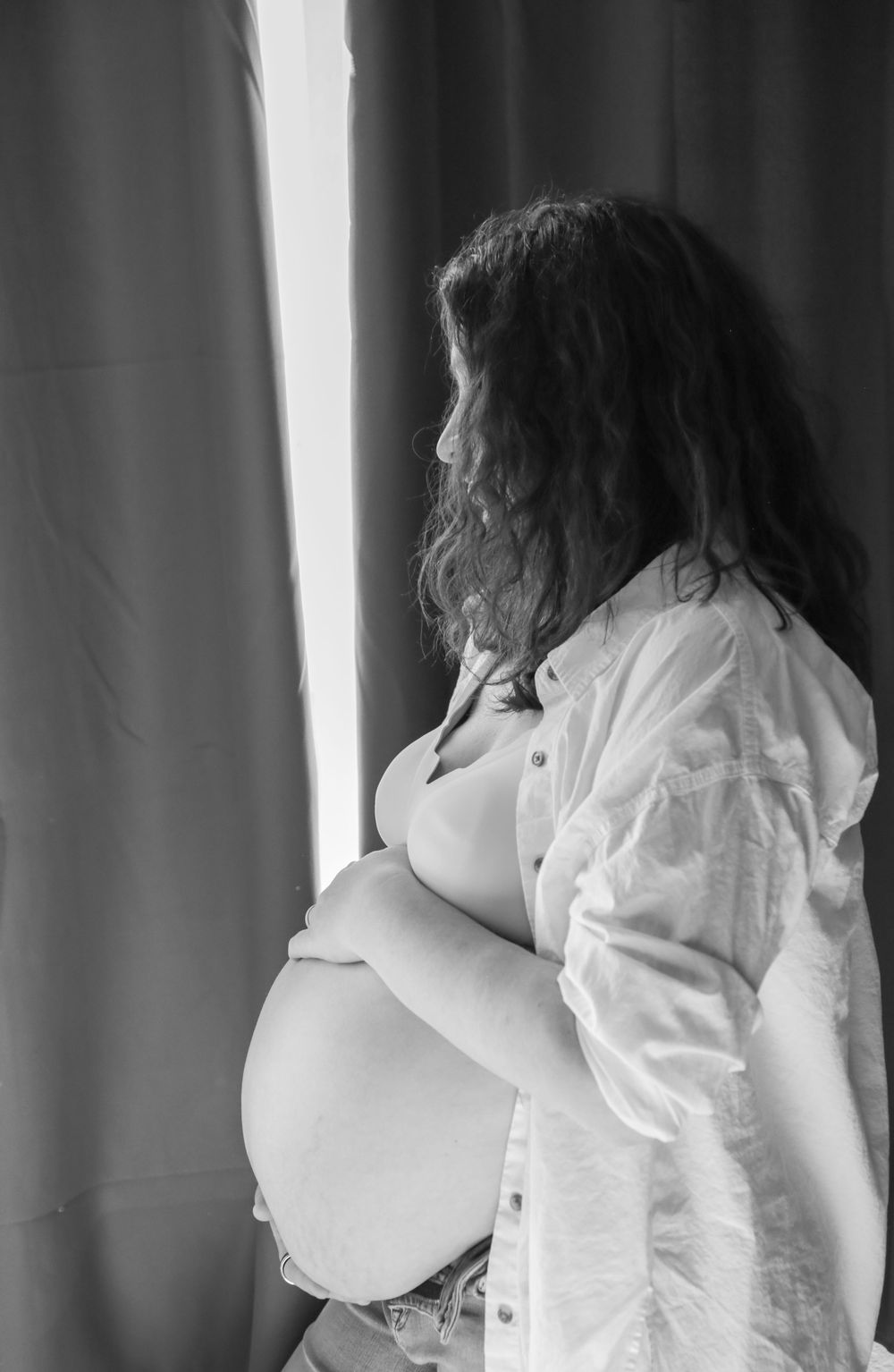 Pregnant woman holding belly standing in front of bedroom window.