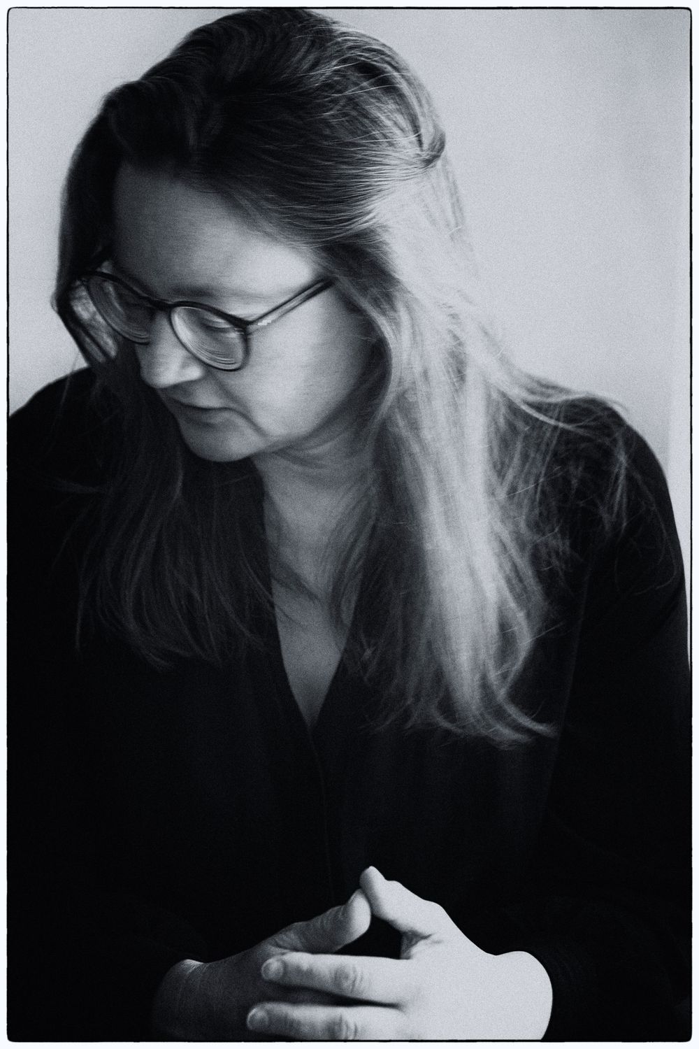 Black and white portrait of a person with glasses and long hair looking down thoughtfully in contemplative pose.