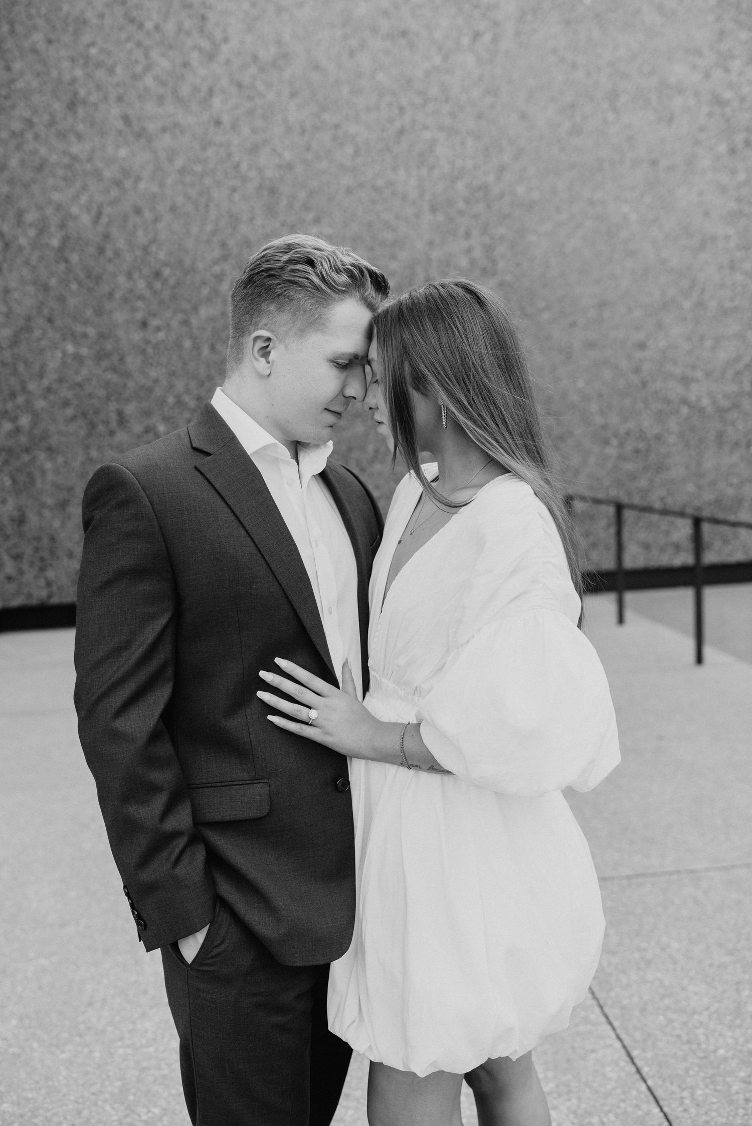 A romantic couple in formal attire share an intimate moment against a textured wall.