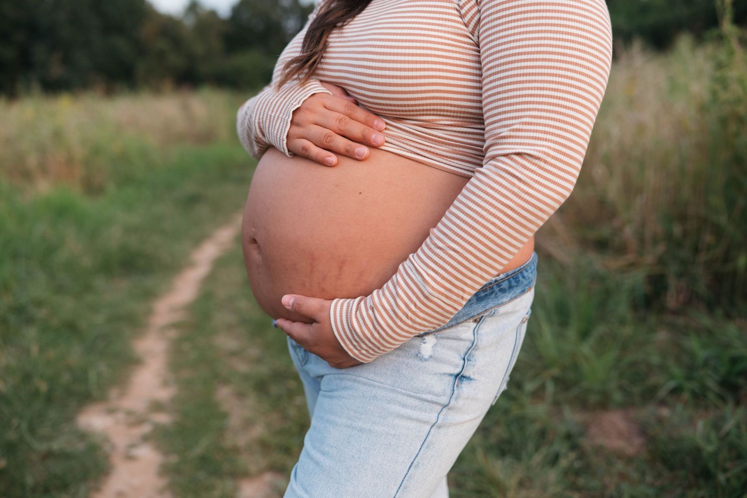 Close up of pregnant belly in striped top against outdoor path.