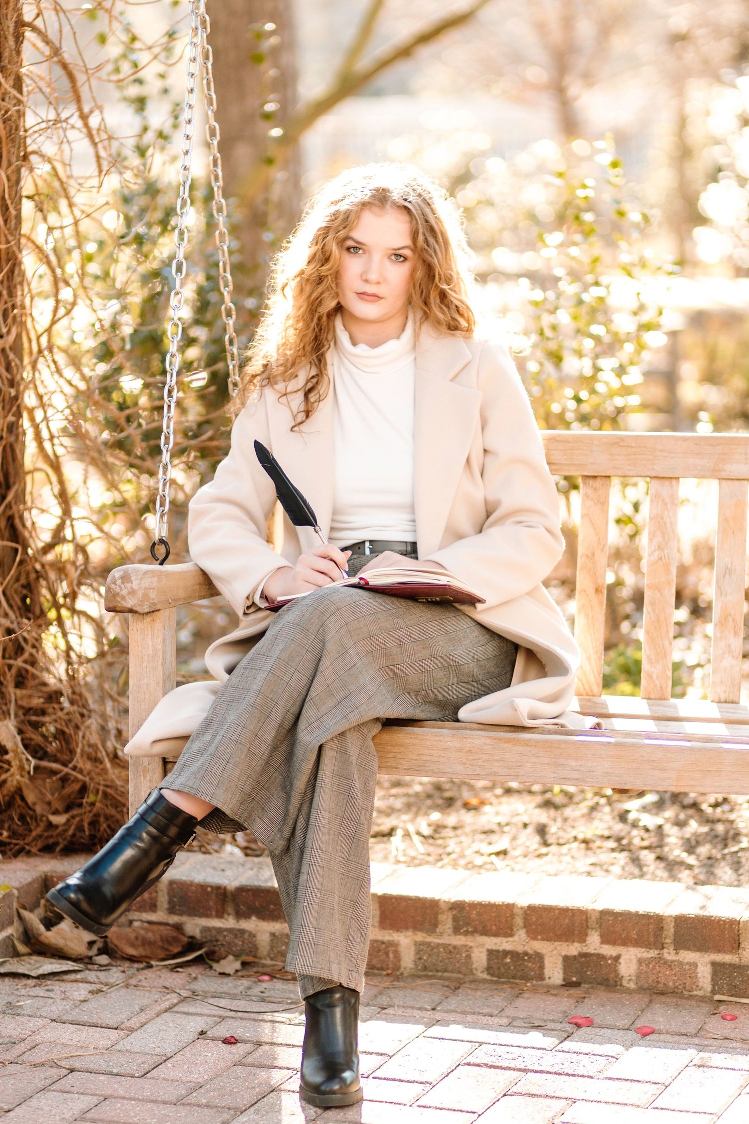 Person in beige coat and grey pants sitting on garden bench writing in notebook on sunny autumn day.