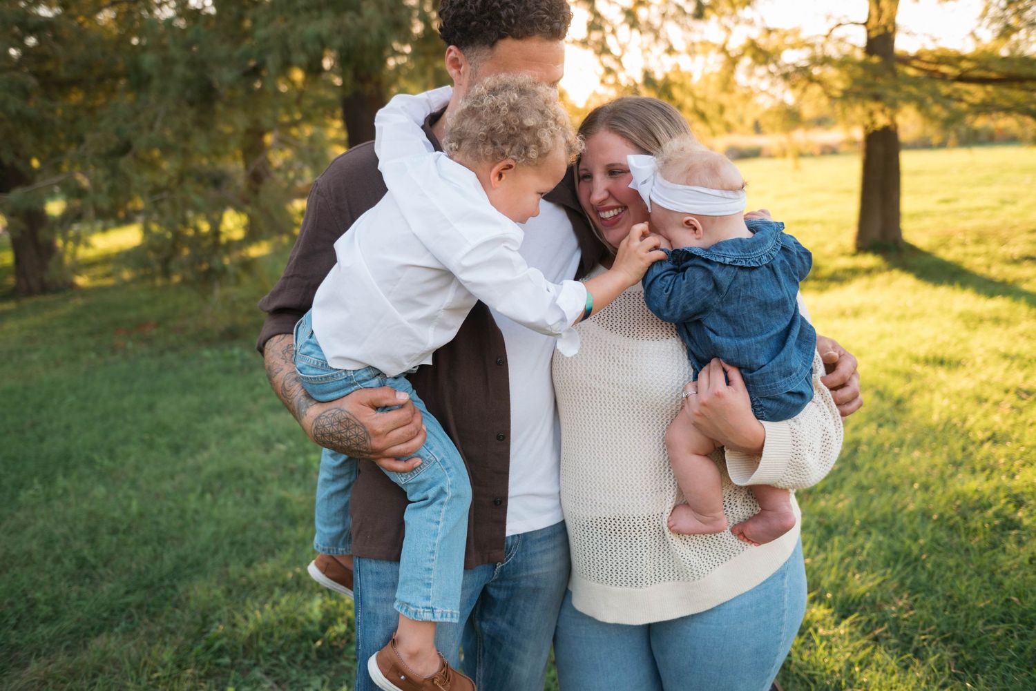 A family wearing coordinated white and denim outfits embraces lovingly in a sunlit park during golden hour.