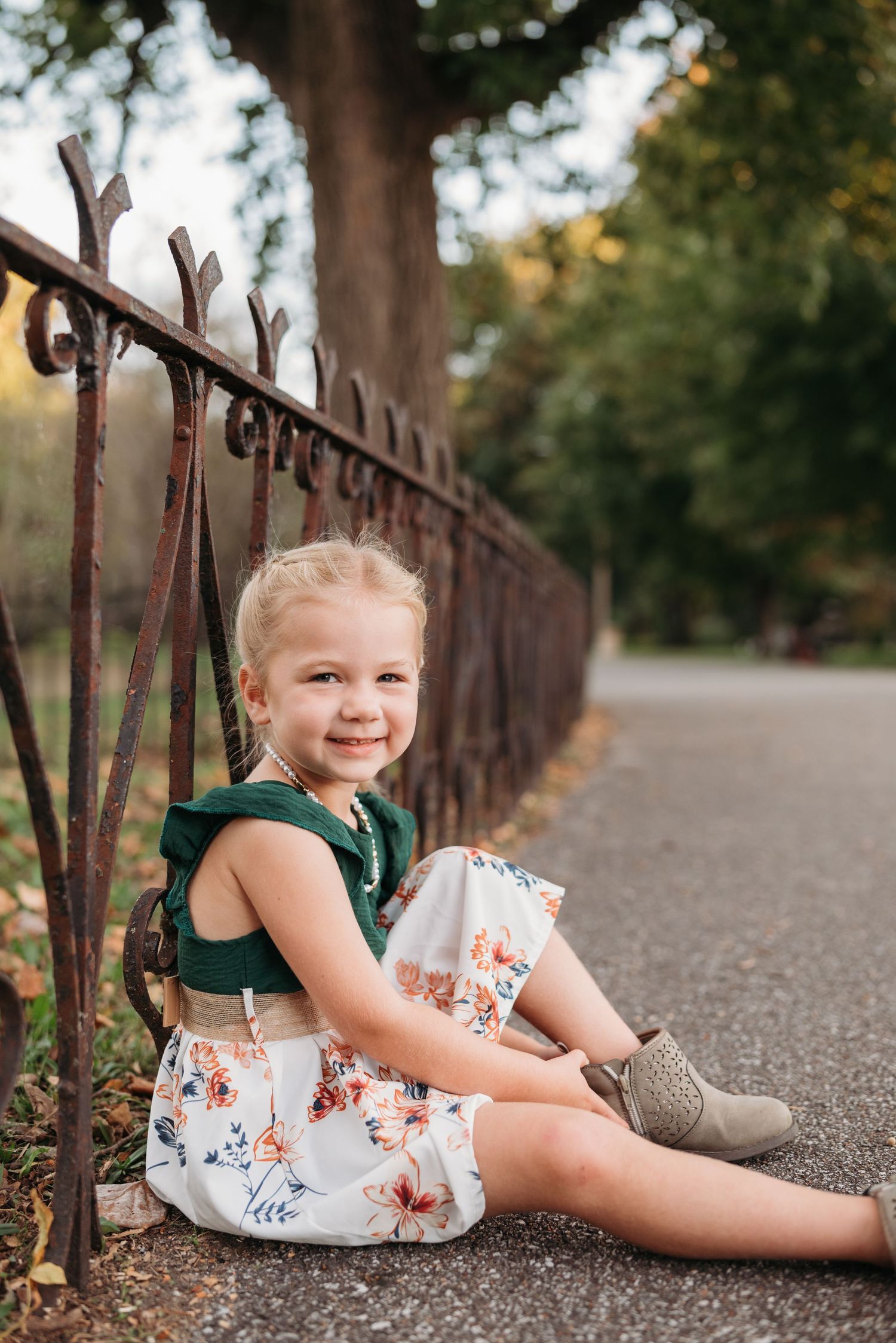 Young child in a floral dress poses playfully by rustic metal railing on a path.