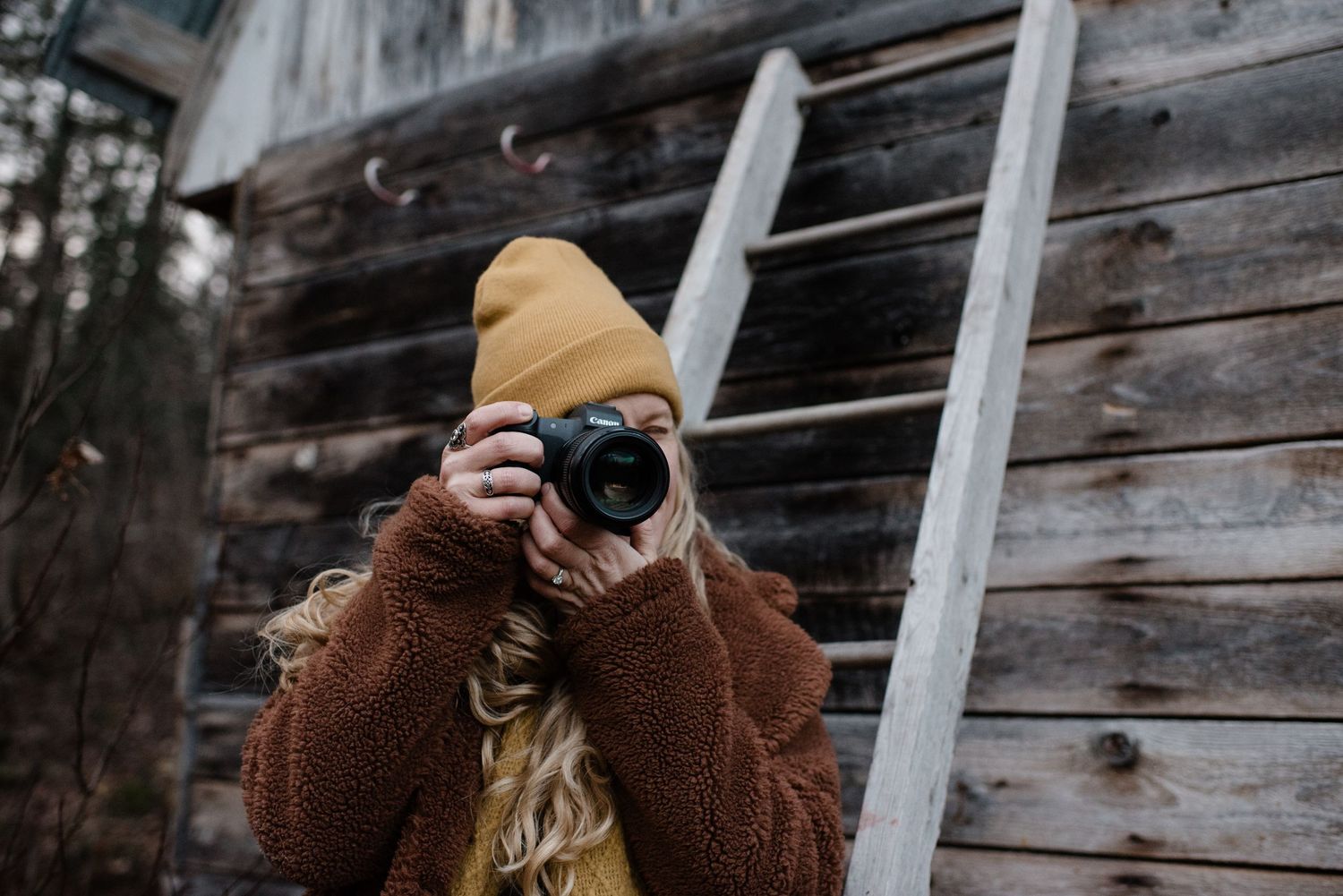 A photographer bundled in a brown fuzzy coat and yellow beanie takes a photo against a rustic wooden wall.