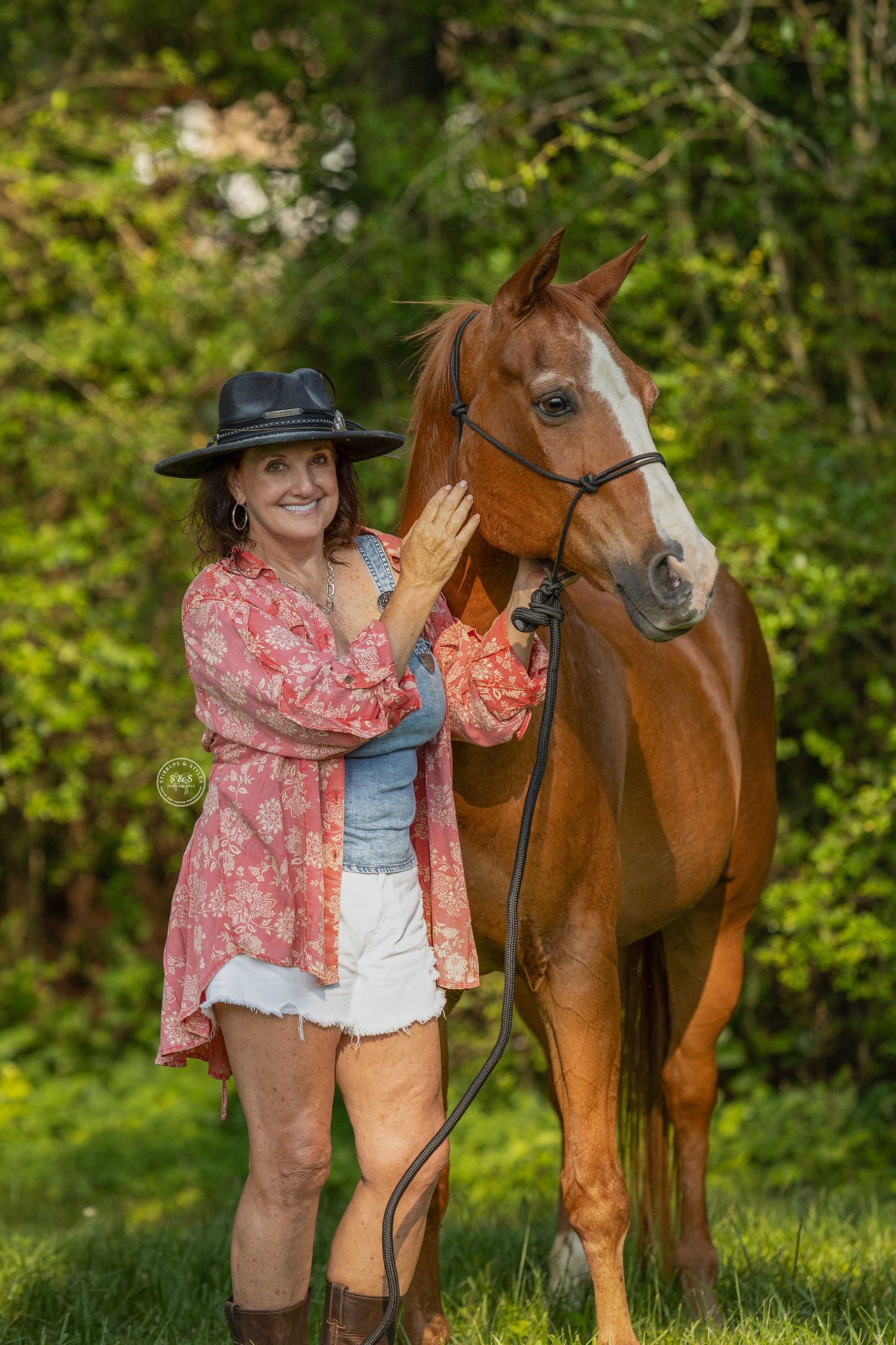 A woman in a pink floral top poses with a chestnut brown horse in a lush green outdoor setting.