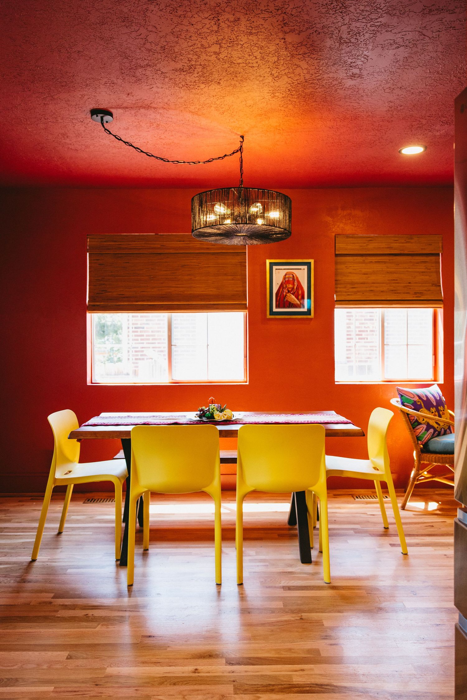 a warm dining room with hardwood floors, orange-red walls, bright yellow chairs, underneath two windows