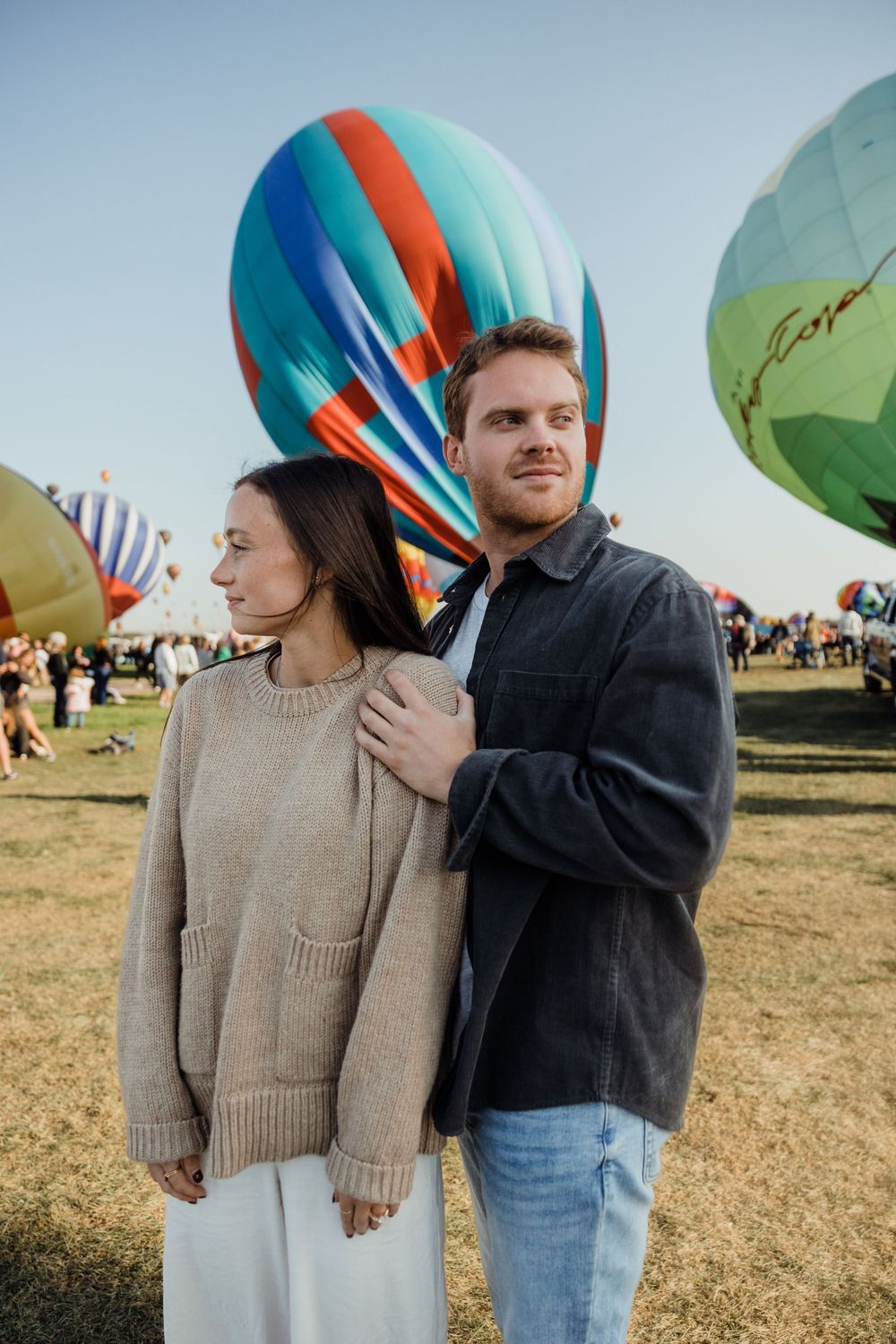 Couple posing at hot air balloon festival, man in dark jacket with arm around woman in beige sweater.