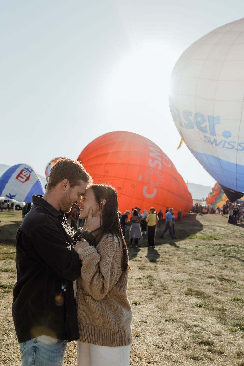Romantic couple embracing face-to-face at sunny hot air balloon festival with colorful balloons behind them.