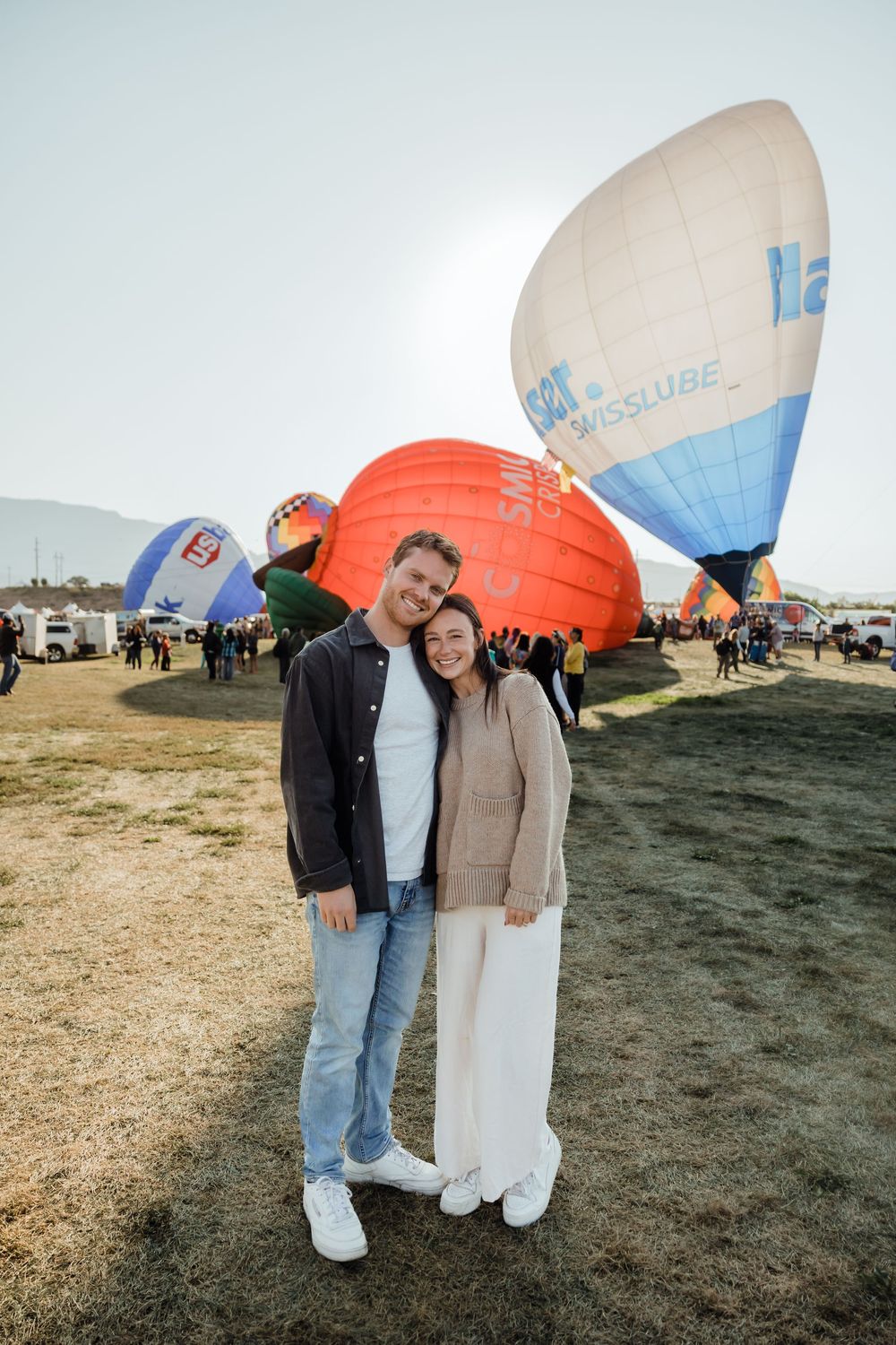 Smiling couple posing together at hot air balloon festival with large orange and white balloons behind them.