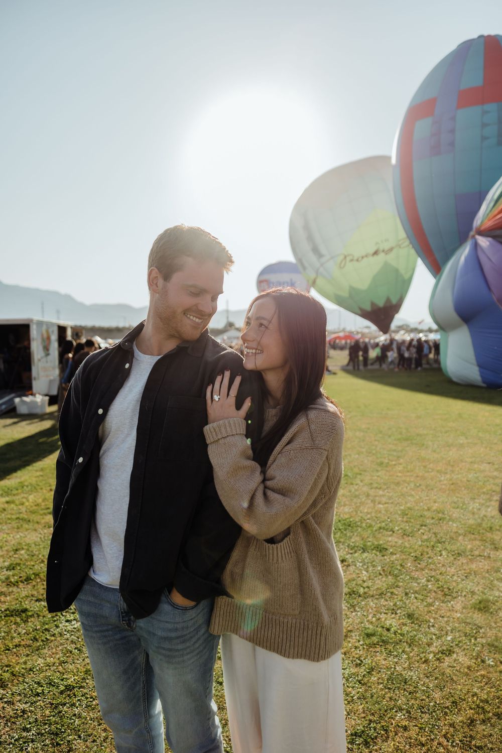 Laughing couple embracing at sunny hot air balloon festival with colorful balloons and mountain backdrop.