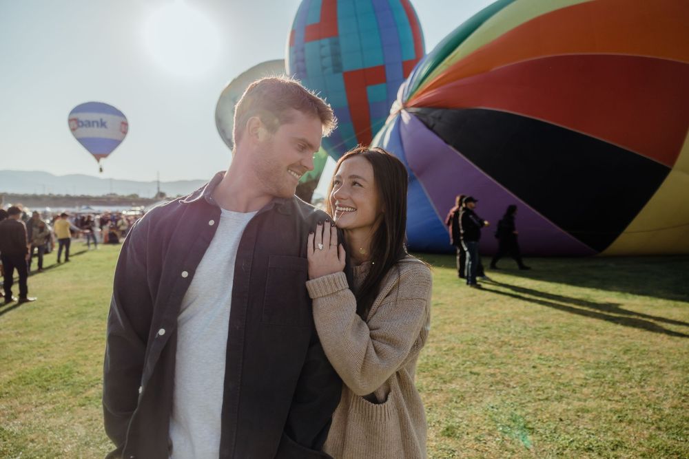 Smiling couple gazing at each other at hot air balloon festival with rainbow-striped balloon behind them.