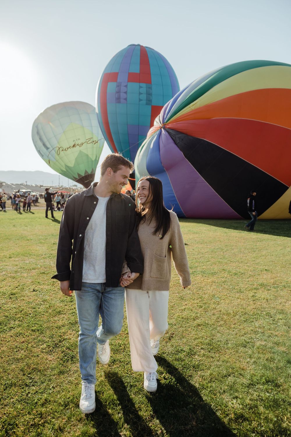A couple holds hands and smiles while walking through a colorful hot air balloon festival on a sunny day.
