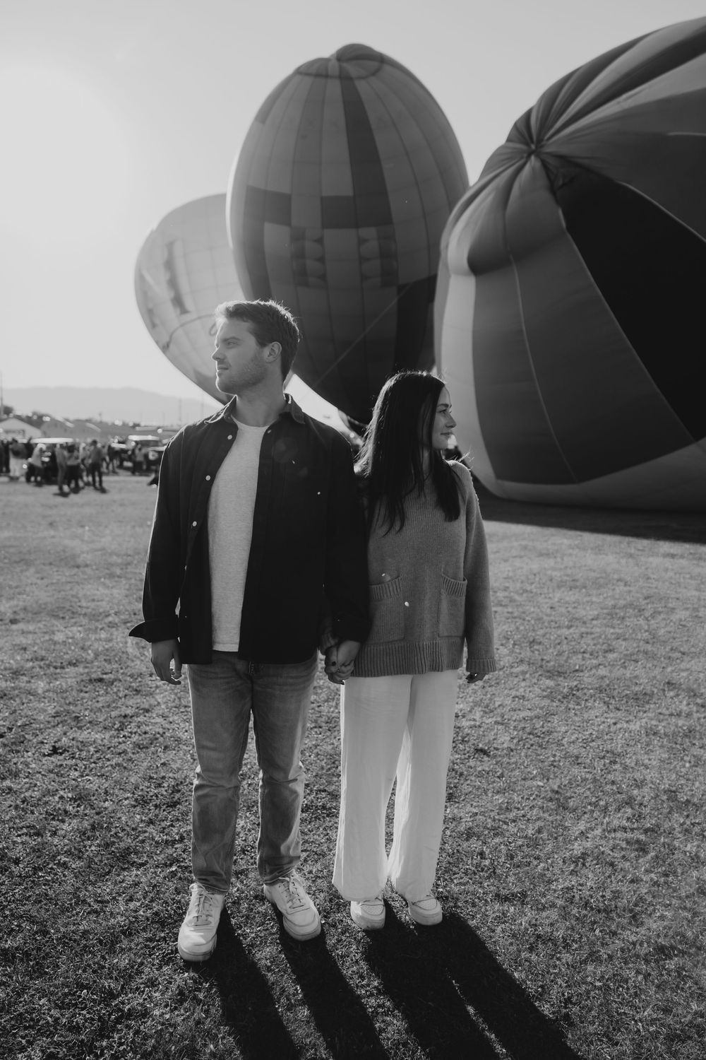 Black and white photo of a couple standing hand in hand surrounded by large hot air balloons on a grassy field.