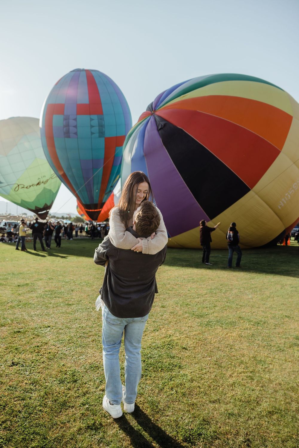 A couple shares a tender embrace on a green field surrounded by colorful inflated hot air balloons.