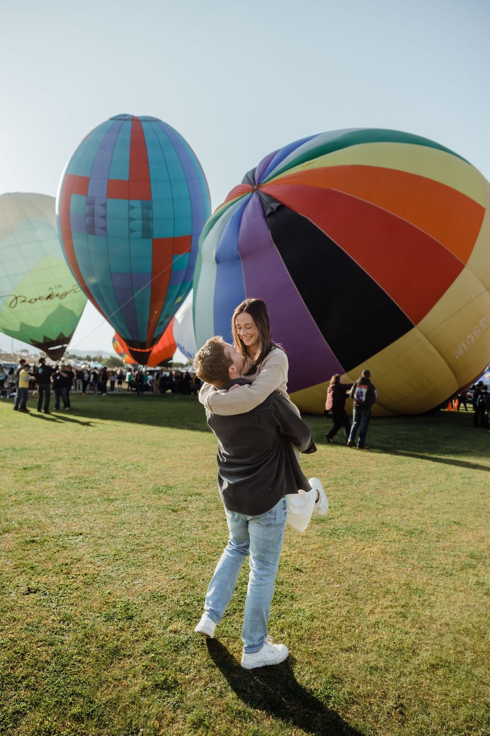 A man lifts and spins a smiling woman at a vibrant outdoor hot air balloon festival on a sunny day.