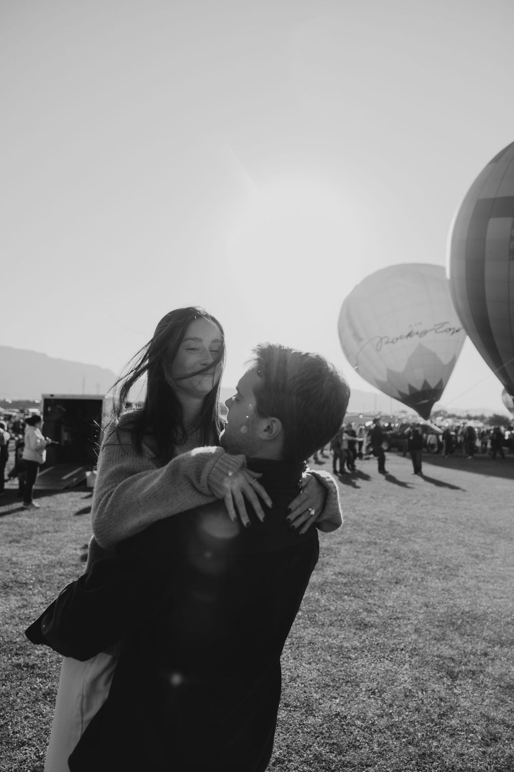 Black and white romantic photo of a man lifting a woman as her hair blows in the wind at a balloon festival.