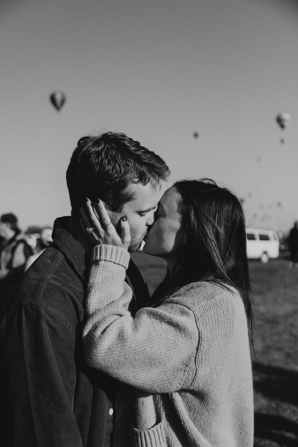Black and white close-up of a couple sharing a kiss with hot air balloons floating in the background.