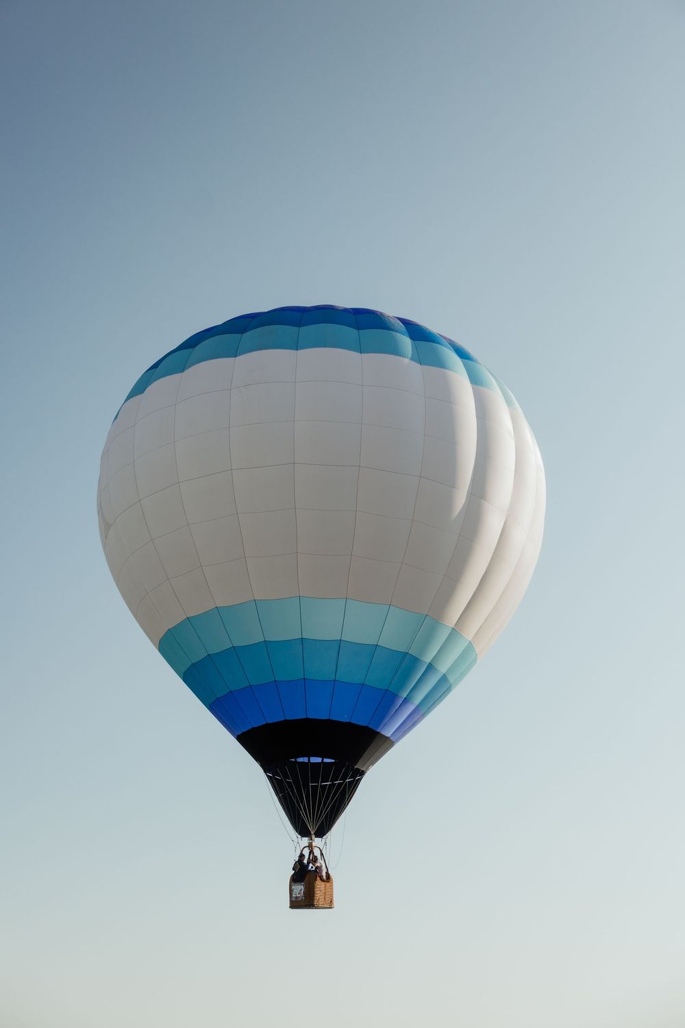 A white and blue striped hot air balloon soars gracefully against a clear pale blue sky.