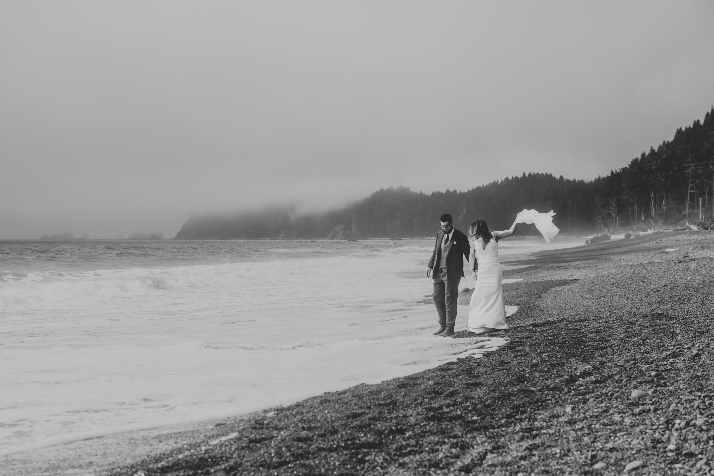 A monochrome image of someone walking along a foggy, rocky beach with hills and houses in the misty distance.