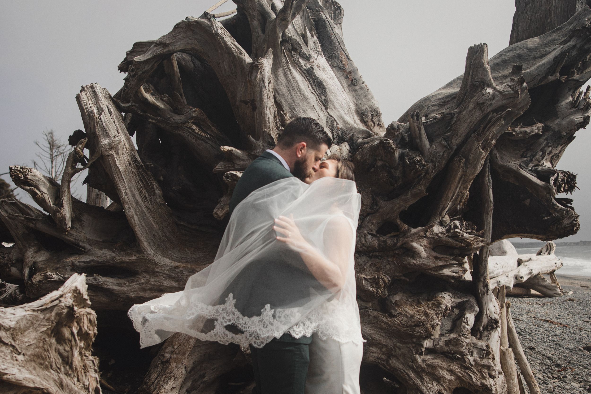 Ethereal white fabric flows in coastal breeze against dramatic driftwood on moody beach landscape.