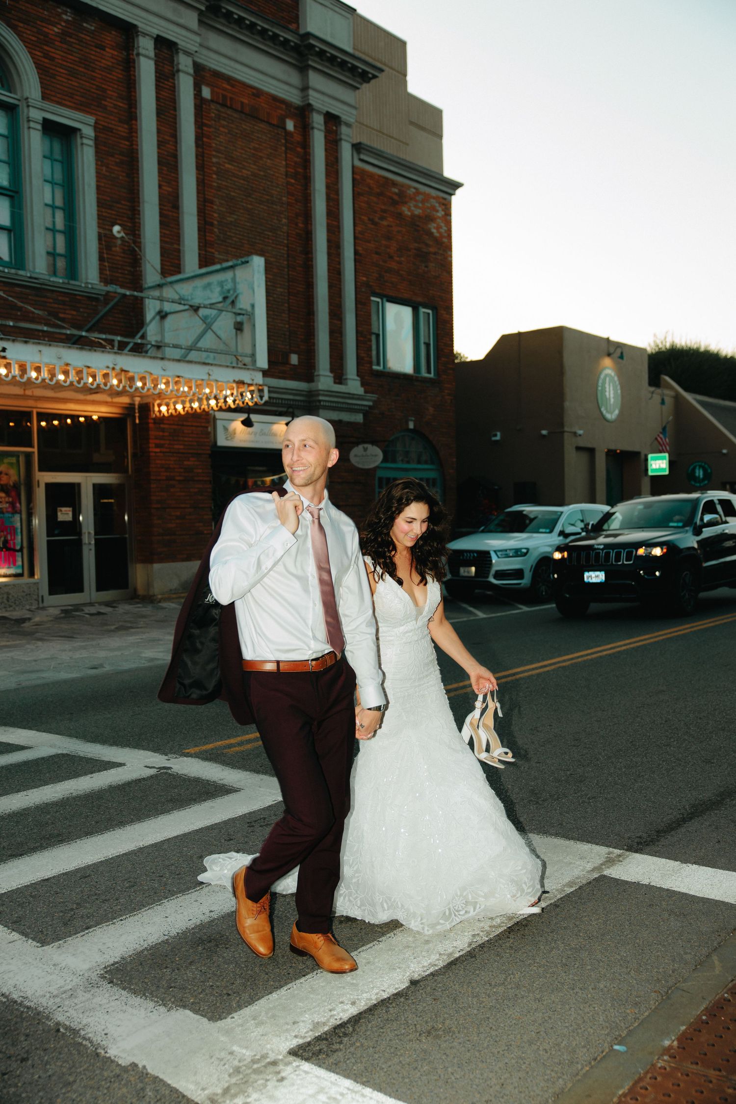 Newlyweds walk across a city street at dusk with brick buildings and string lights in the background.