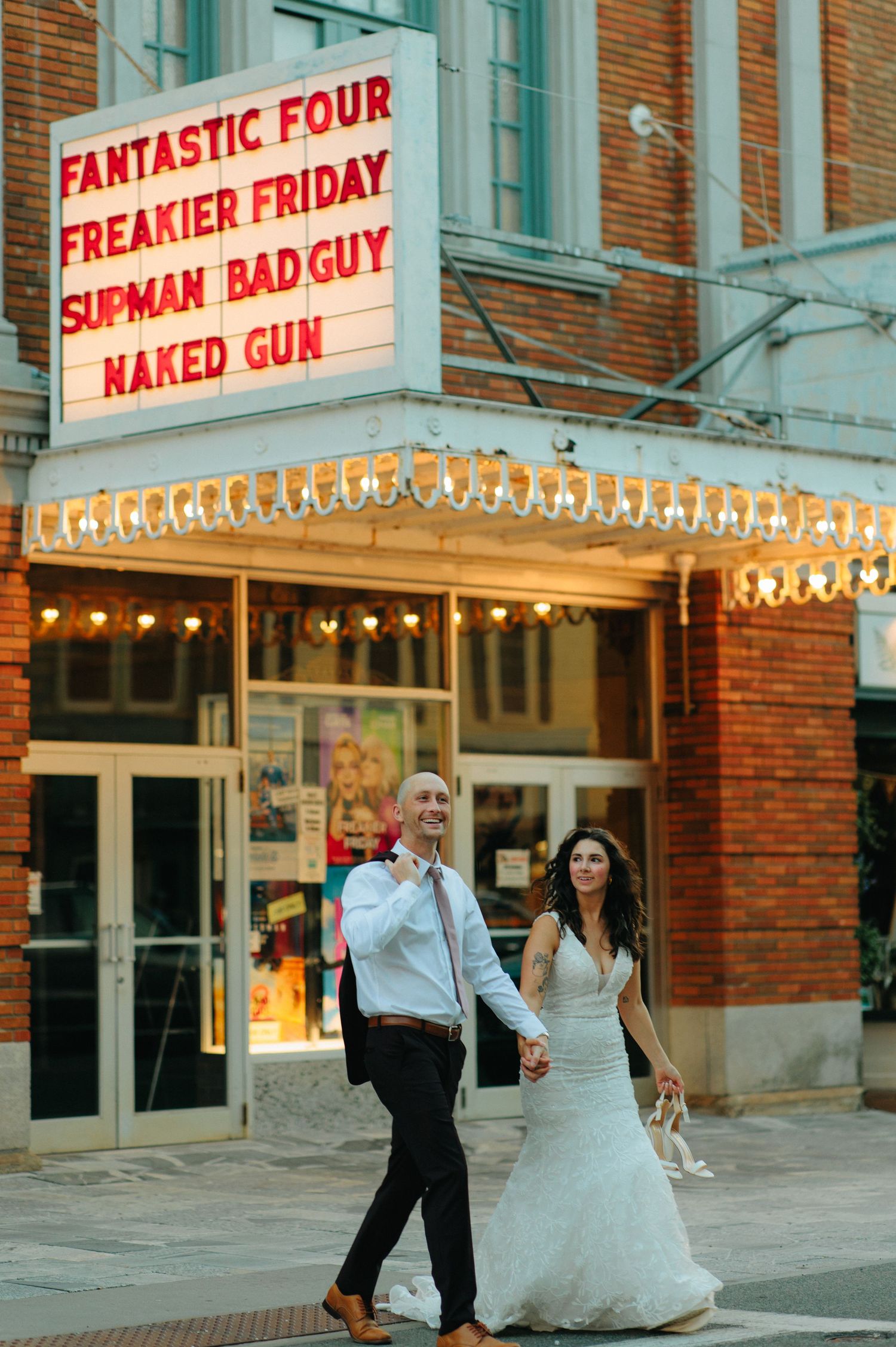 Wedding couple walking past vintage movie theater marquee with glowing lights at dusk.