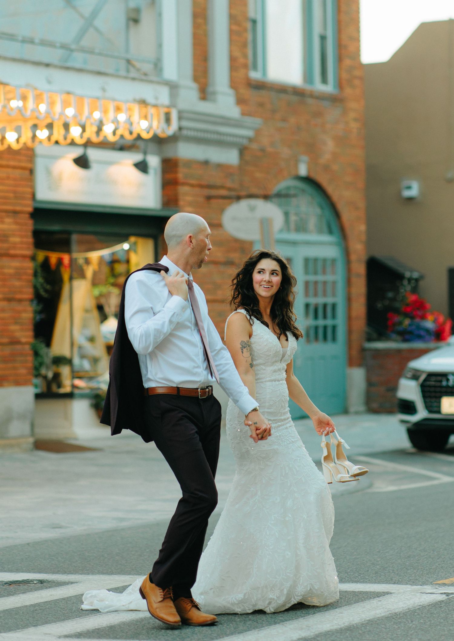 A couple in wedding attire walks confidently across a city street at dusk with string lights and brick buildings behind them.