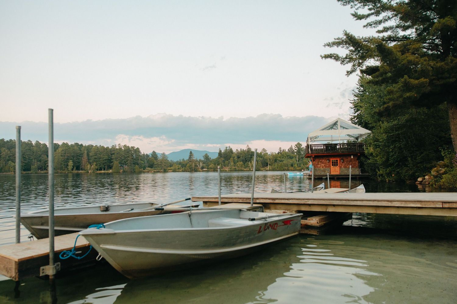 A boat dock stretches over calm lake waters at dusk with a forested shoreline and cloudy sky in the background.