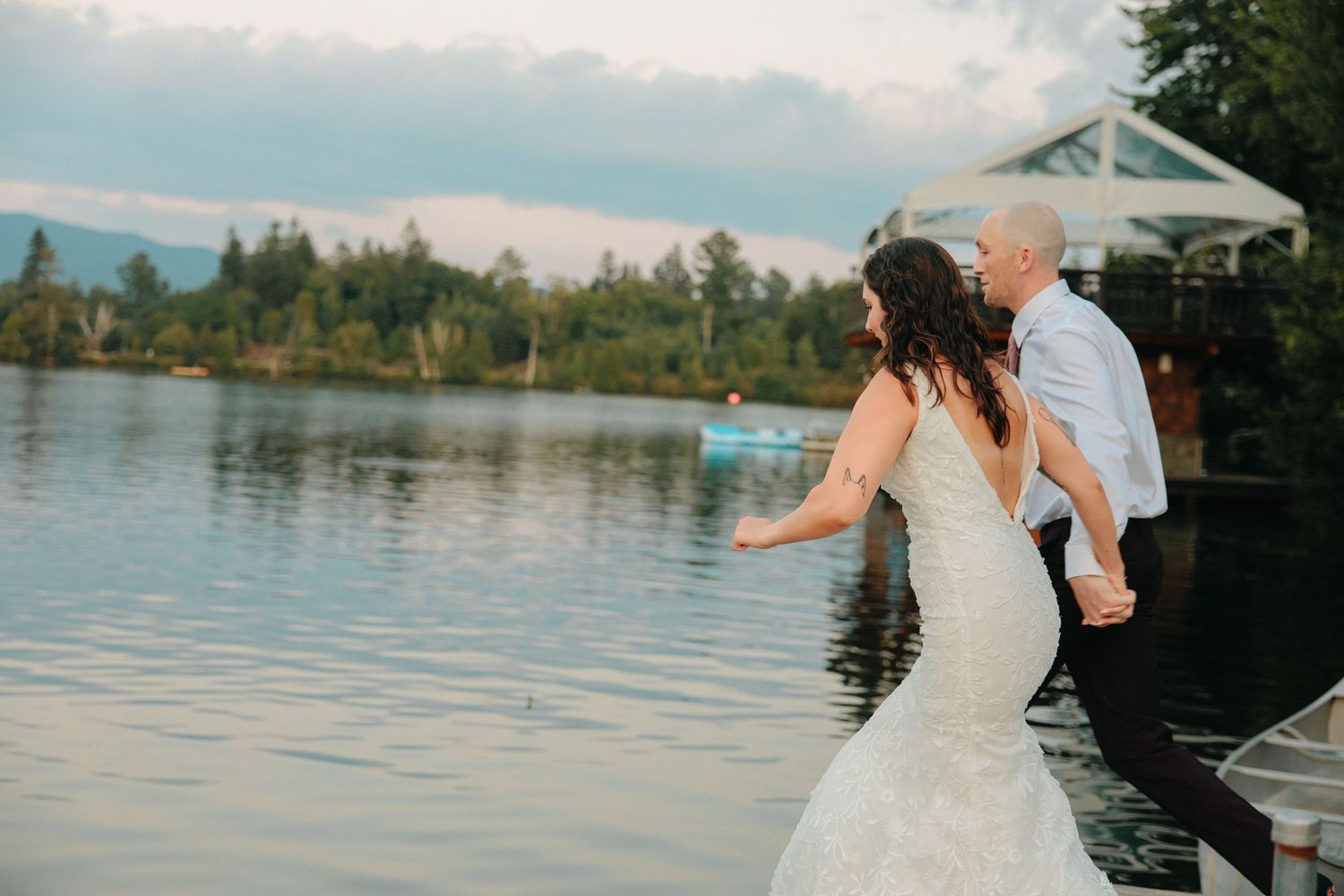 A couple in wedding attire share a romantic moment on a lakeside dock at sunset.