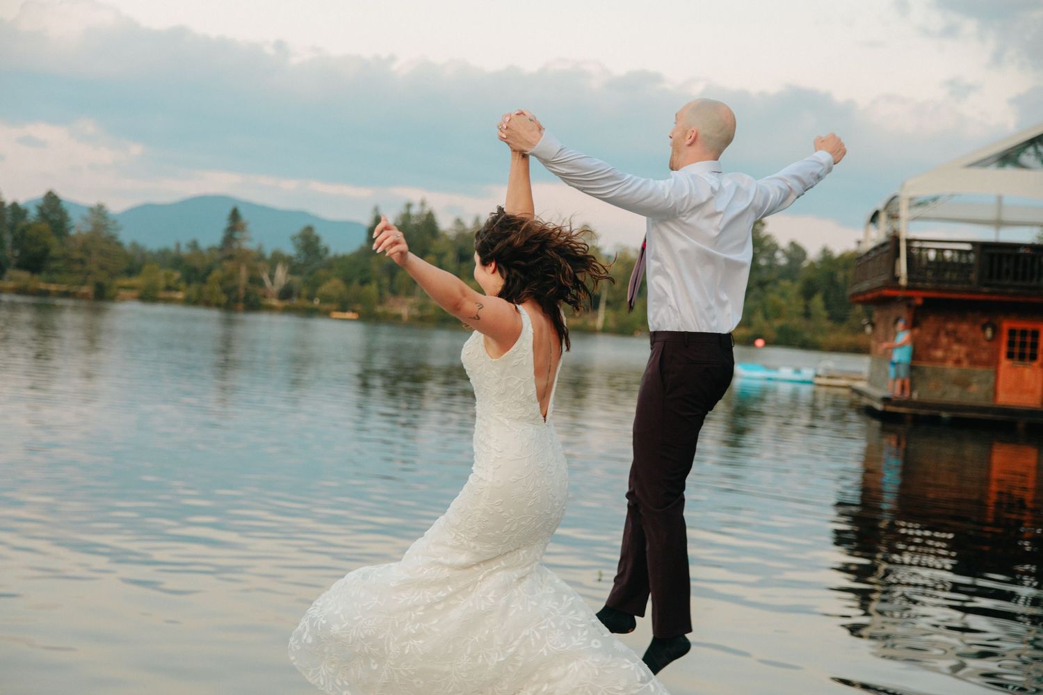 Newlyweds joyfully jumping together by a scenic lake with mountains in the background.