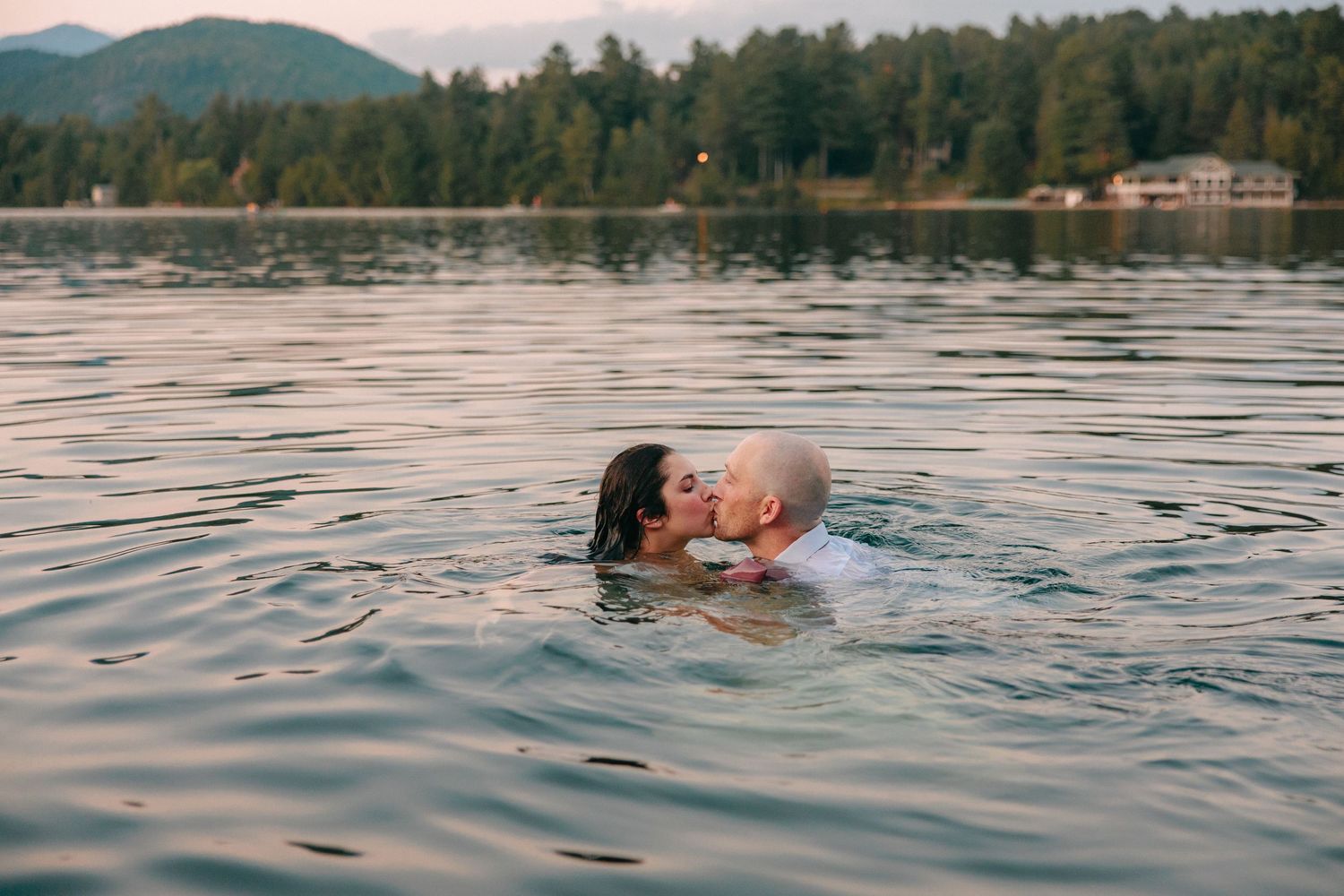 Couple swimming in a serene lake with mountains and trees in the distance.