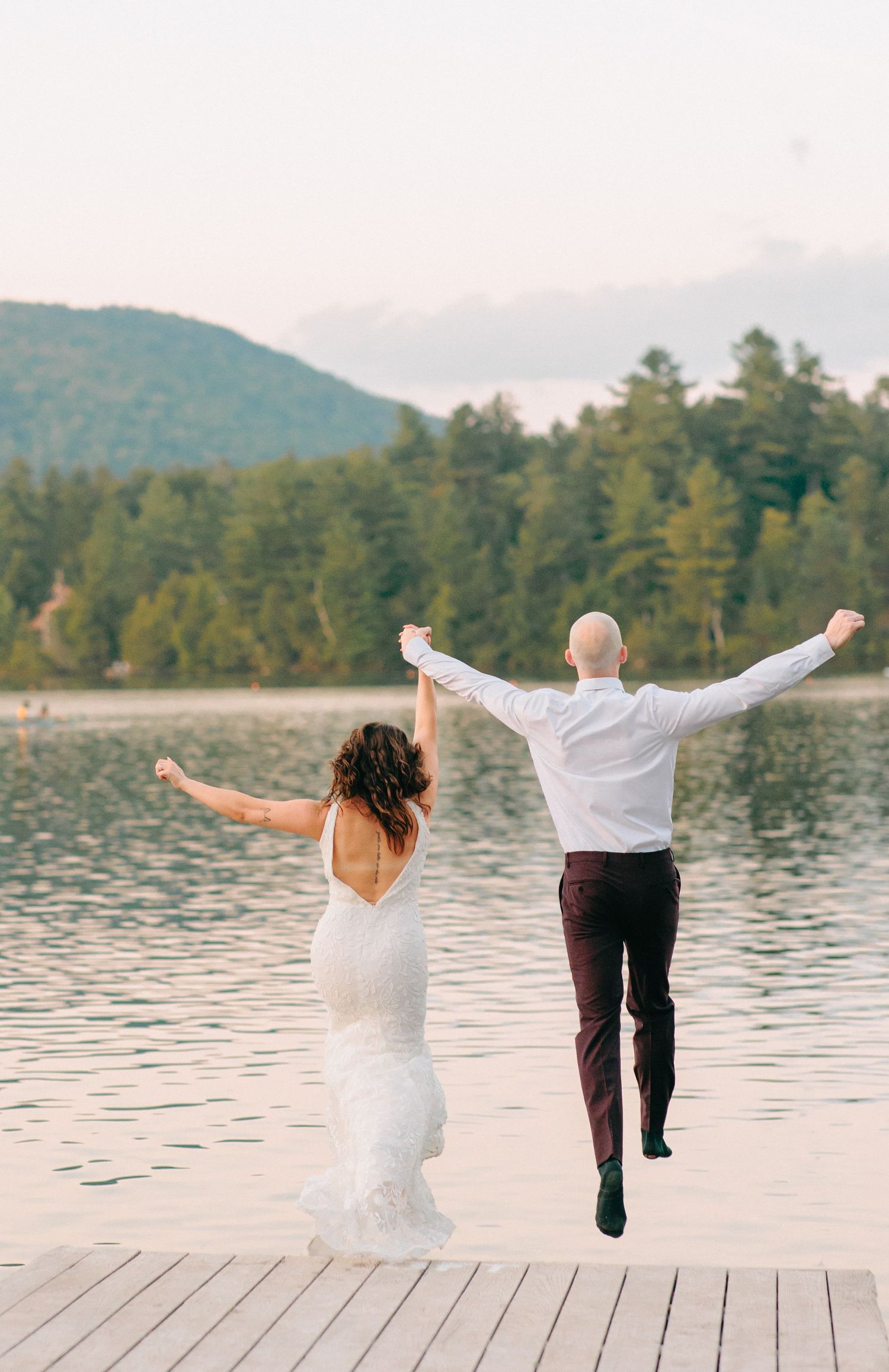 Two people in wedding attire jump joyfully off a dock into a lake with mountains and forest in the background.