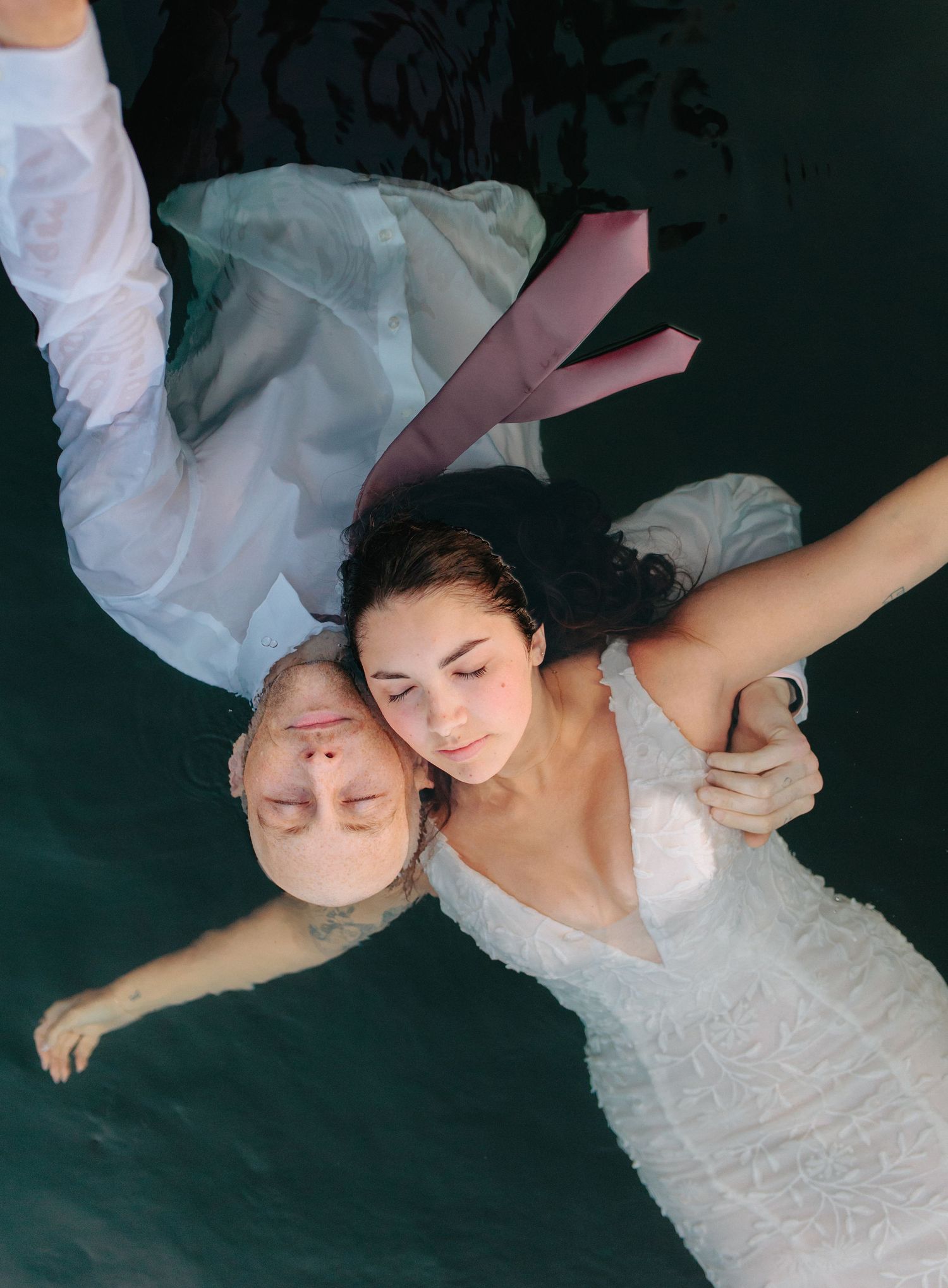 Wedding couple lying together in artistic overhead shot with white attire against dark background.