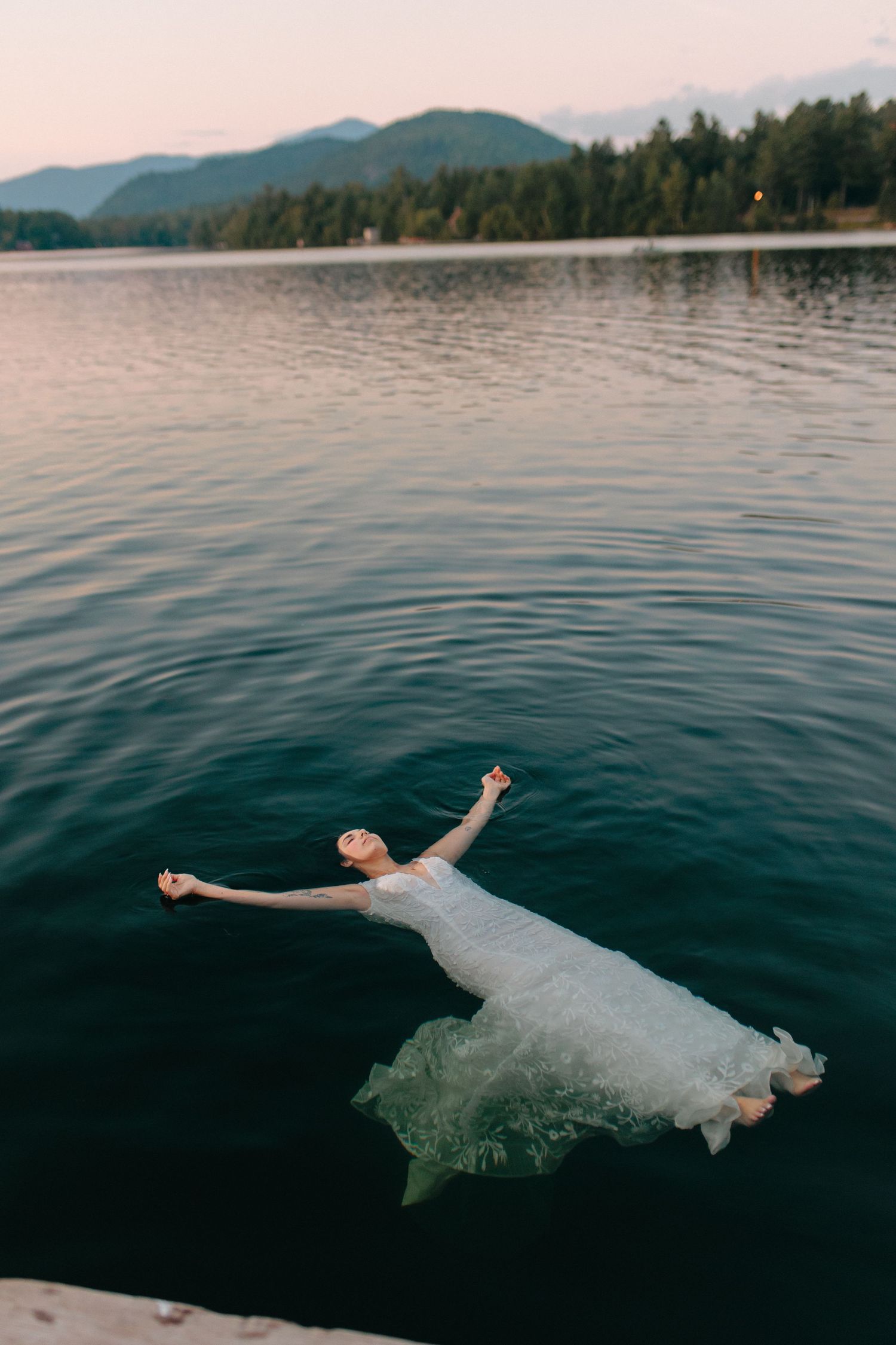 A bride in a flowing white dress floats gracefully in dark lake waters with mountains in the background.