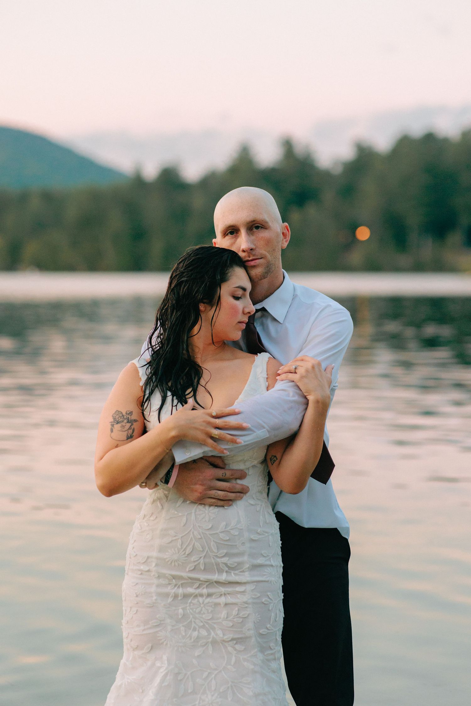 Romantic couple embraces by a serene lake at sunset with mountains in background.