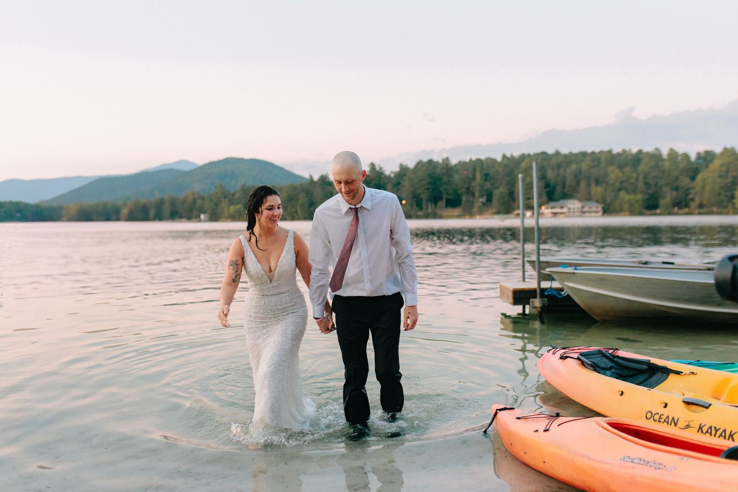 Couple wading in lake water during sunset with boats and kayaks on shoreline in background.