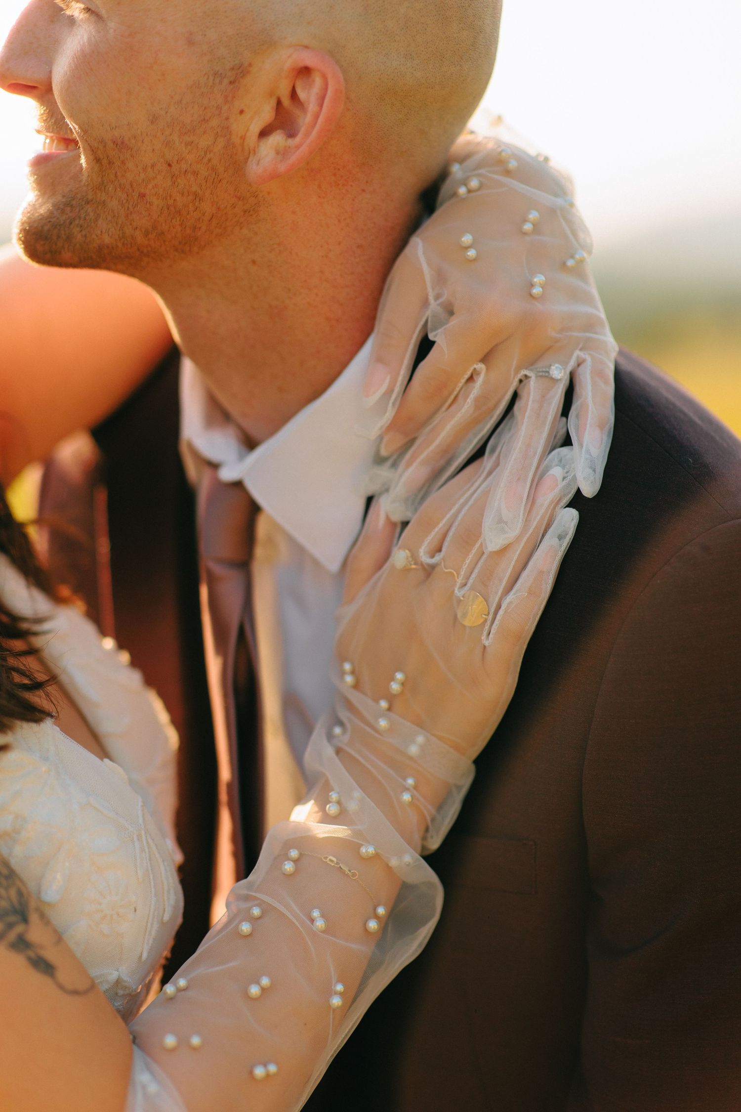 Close-up of elegant bridal glove with pearl details resting against a formal suit at sunset.