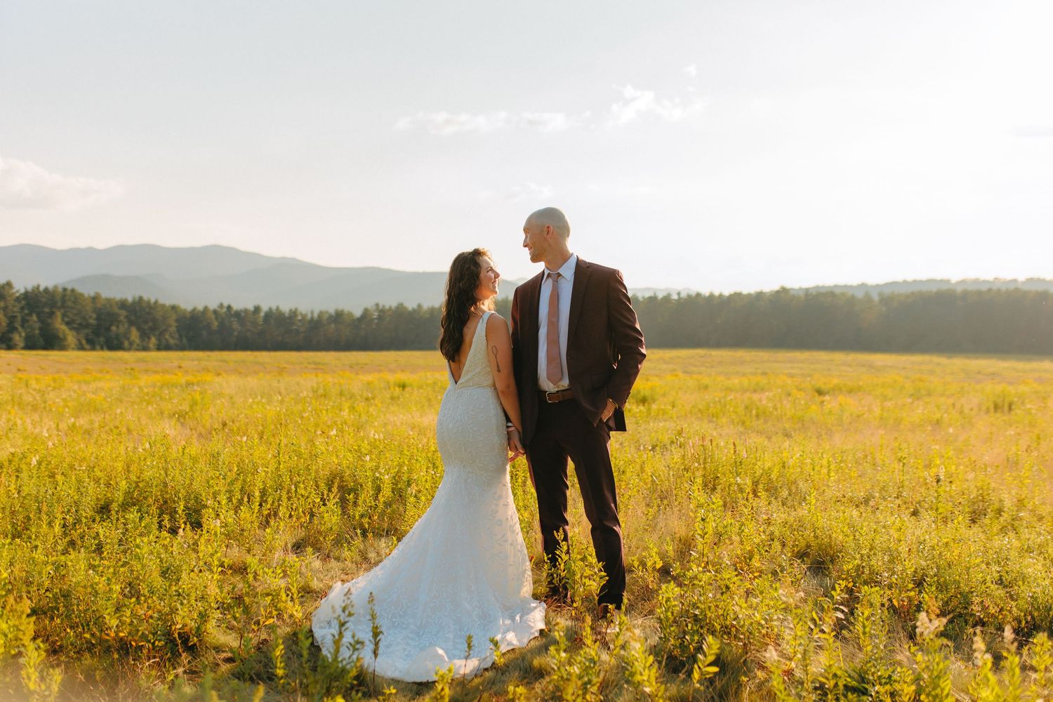 A couple shares an intimate moment in a golden meadow with mountains in the background during sunset.