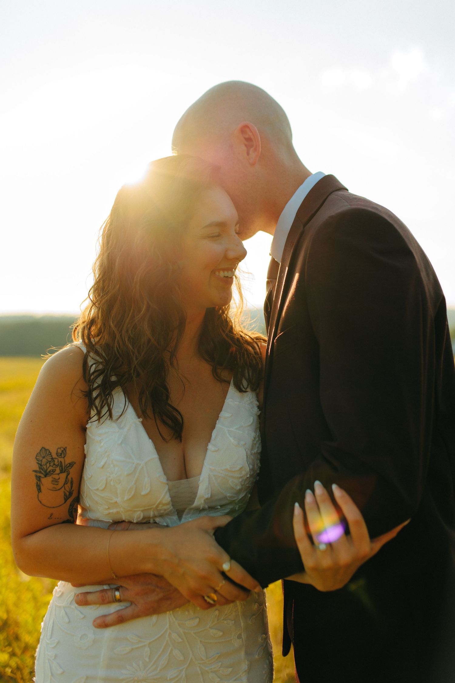 A romantic wedding couple embraces in a sunlit field during golden hour with a bride in white lace gown.
