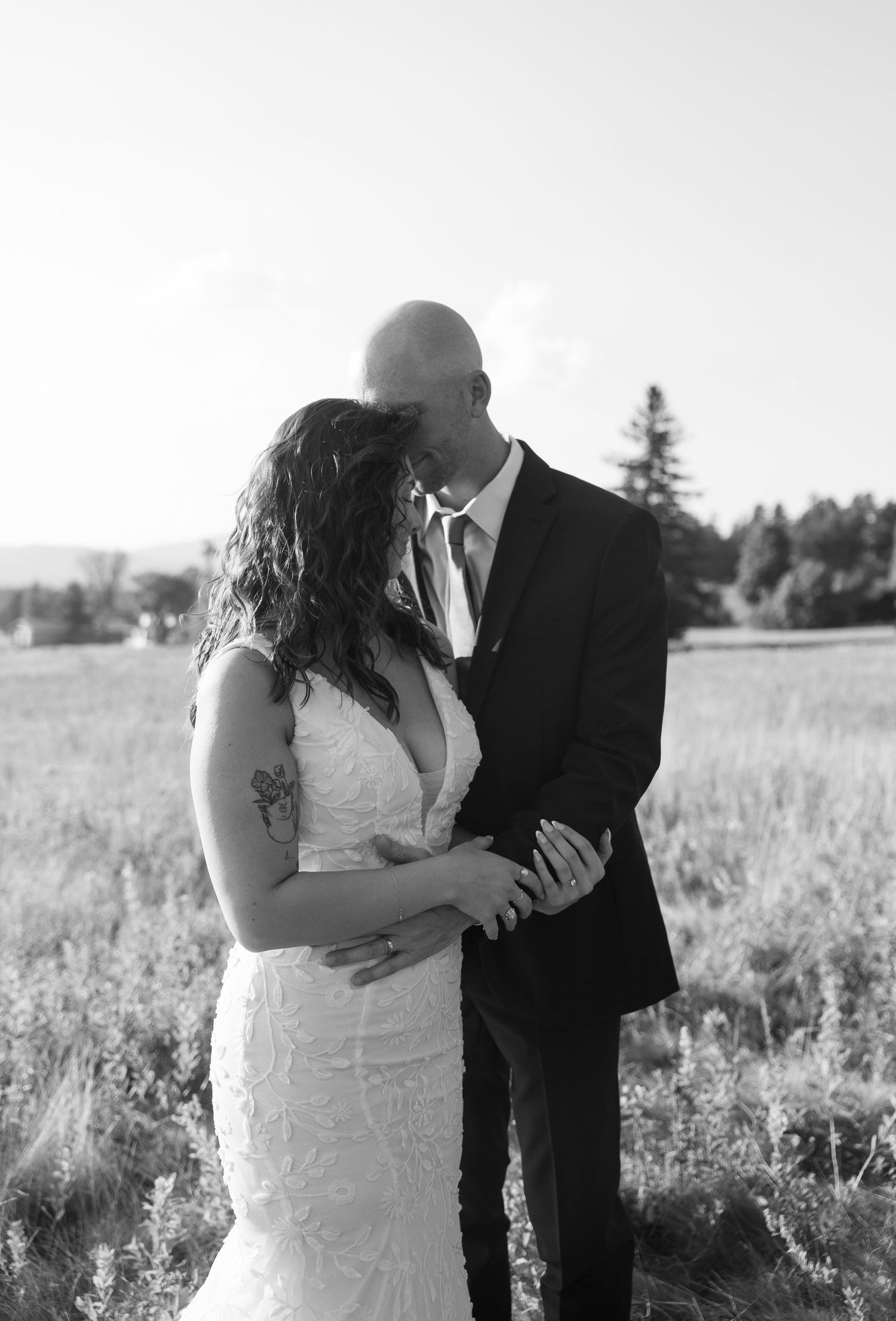 A romantic black and white wedding photo showing a couple embracing in a field with trees in the background.