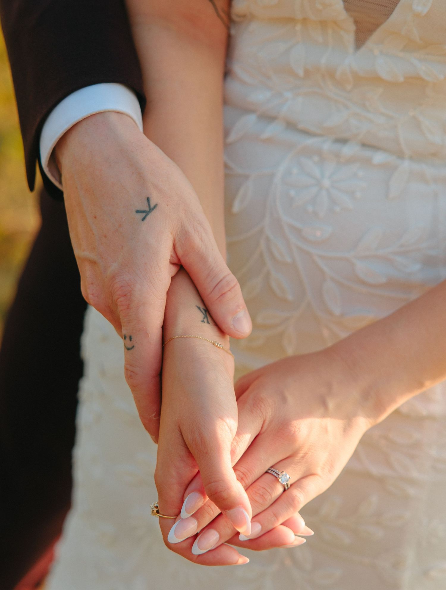 A closeup of clasped hands showing wedding rings and small cross tattoos during a wedding ceremony.