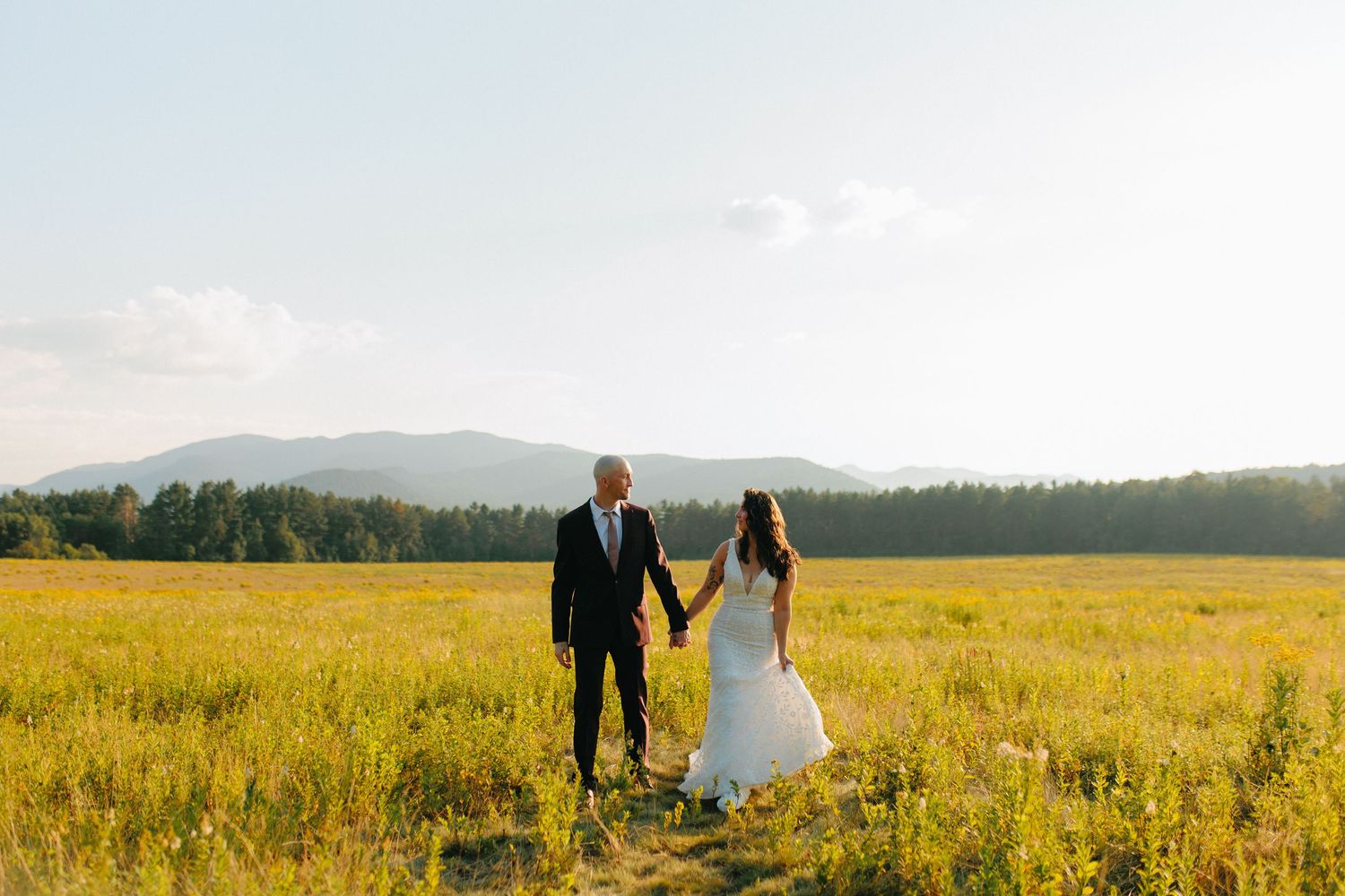 Wedding couple embraces in tall grass meadow with mountains and forest in the distance at golden hour.